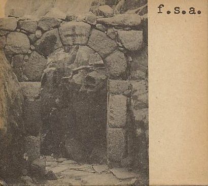 Photo of a rudimentary stone archway, potentially medieval, with a cross engraving on the top middle of the arch. The photo is in black and white, printed on a brownish cardboard, making it look rugged, mysterious and eerie